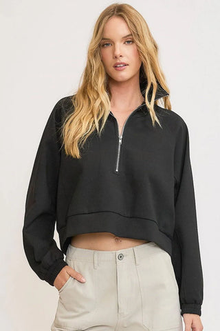 Woman with long blonde hair wearing a black cropped zip-up sweatshirt and beige pants, standing against a light gray background with one hand in po...