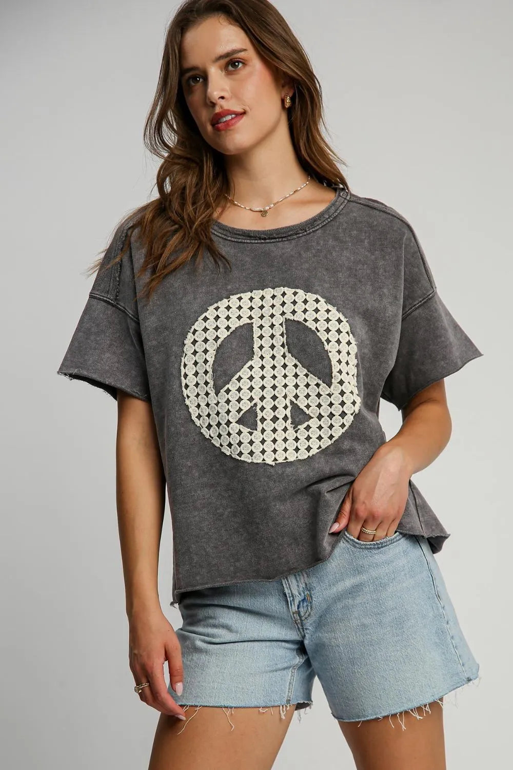 Woman wearing gray vintage-style oversized t-shirt with white checkered peace sign graphic, light blue denim shorts, and layered necklaces against ...