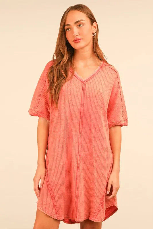 Woman with long brown hair wearing a coral pink ribbed v-neck dress with short sleeves, posed against a cream background.