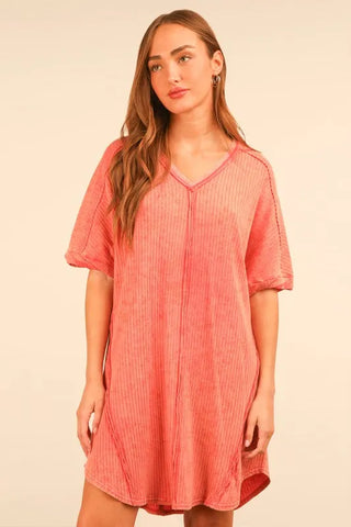 Woman with long brown hair wearing a coral pink ribbed v-neck dress with short sleeves, posed against a cream background.