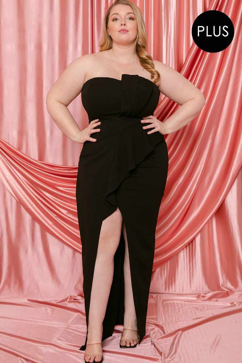 Plus size black pleated maxi dress with tube top, front ruffle, and slit, modeled in front of pink draped backdrop.