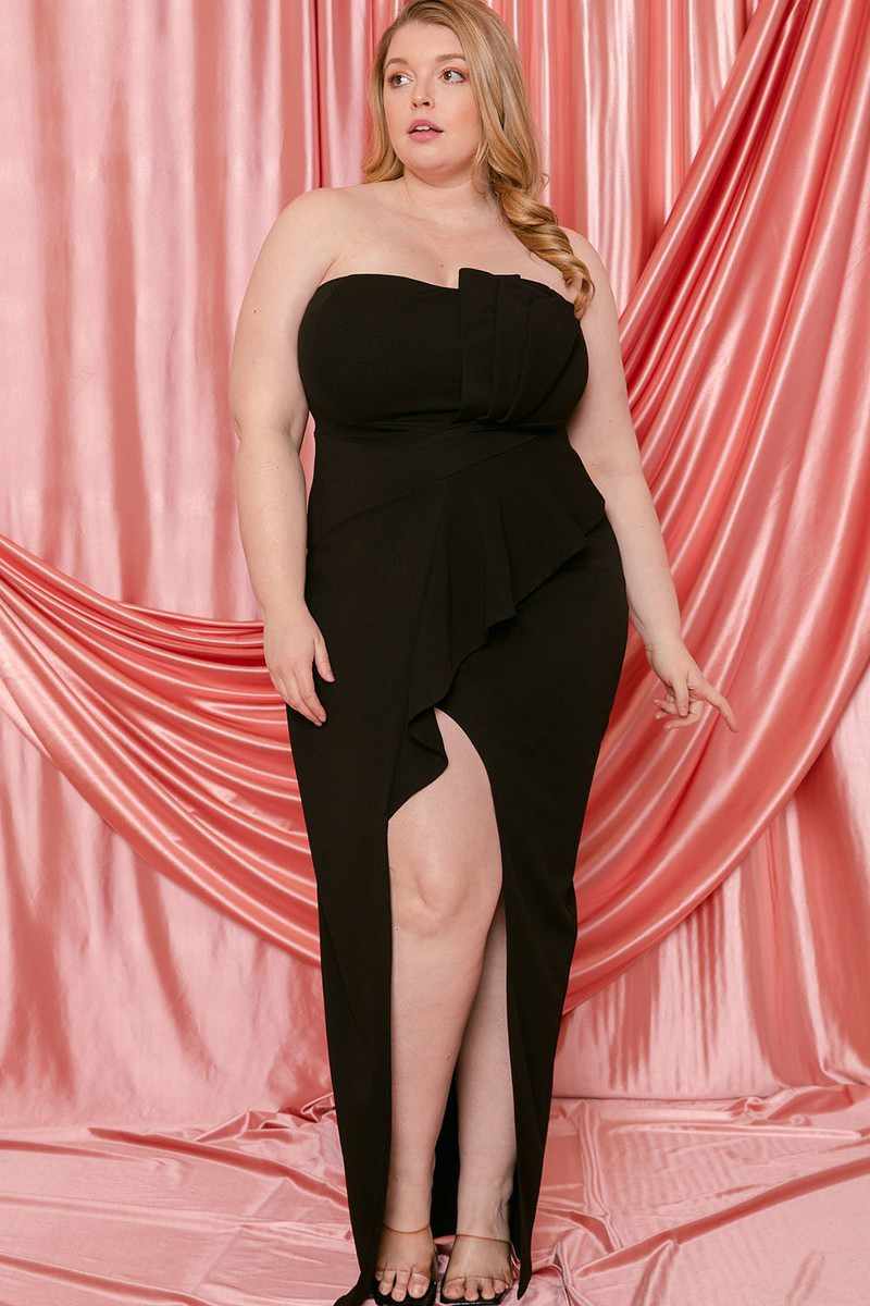 Plus size black pleated tube top maxi dress with ruffle and front slit, featuring back zipper, displayed against pink satin curtain.