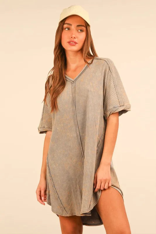 Woman wearing gray ribbed oversized t-shirt dress and white baseball cap, posing against cream background with warm lighting. She has long brown ha...