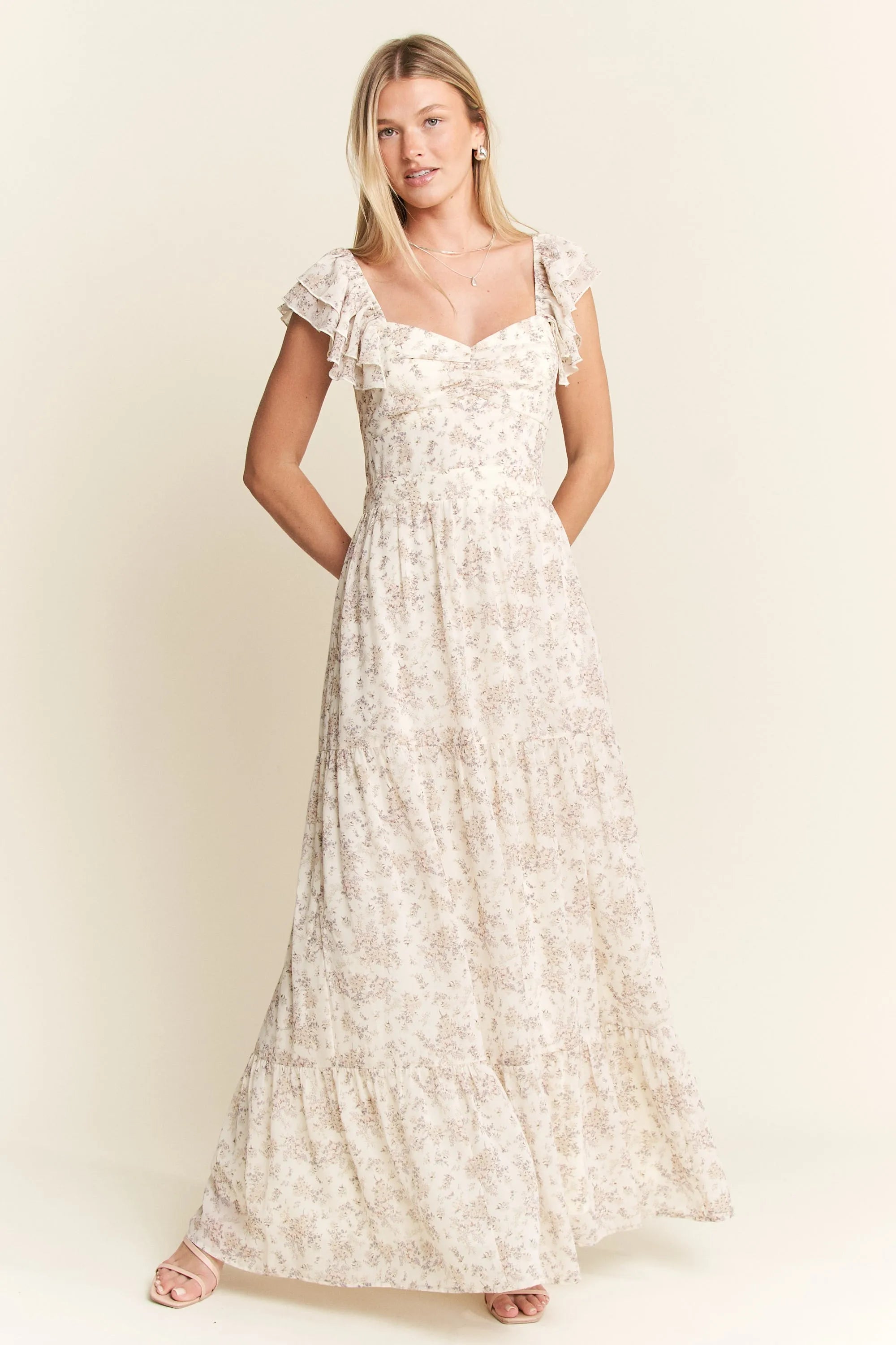 Woman in cream-colored floral maxi dress with ruffled sleeves and tiered skirt, standing with hands on hips against beige background. She has blond...