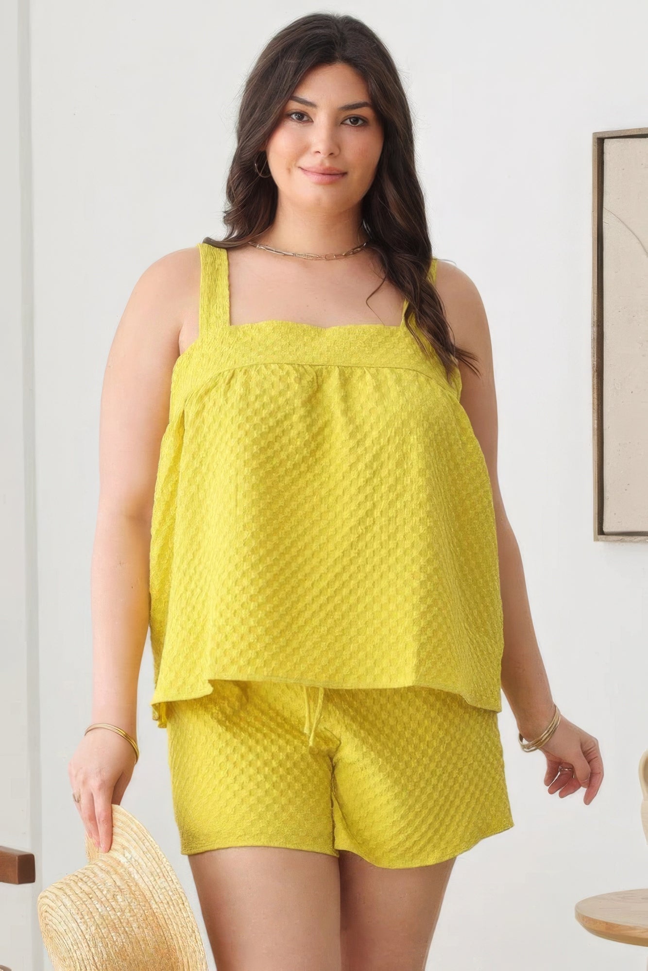 Woman wearing plus size sleeveless textured top and elastic waist shorts in dijon color, holding a hat, showcasing spring fashion.