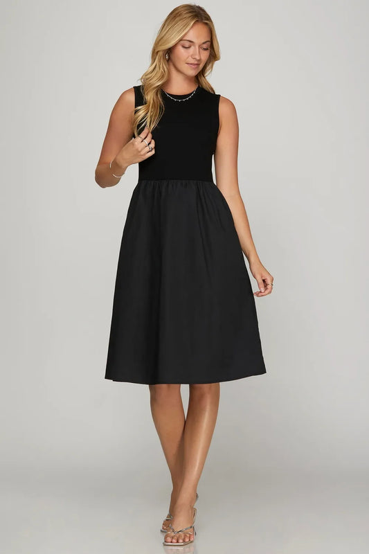 Woman in black sleeveless midi dress with fitted top and A-line skirt, wearing strappy sandals and delicate jewelry, standing against light gray ba...