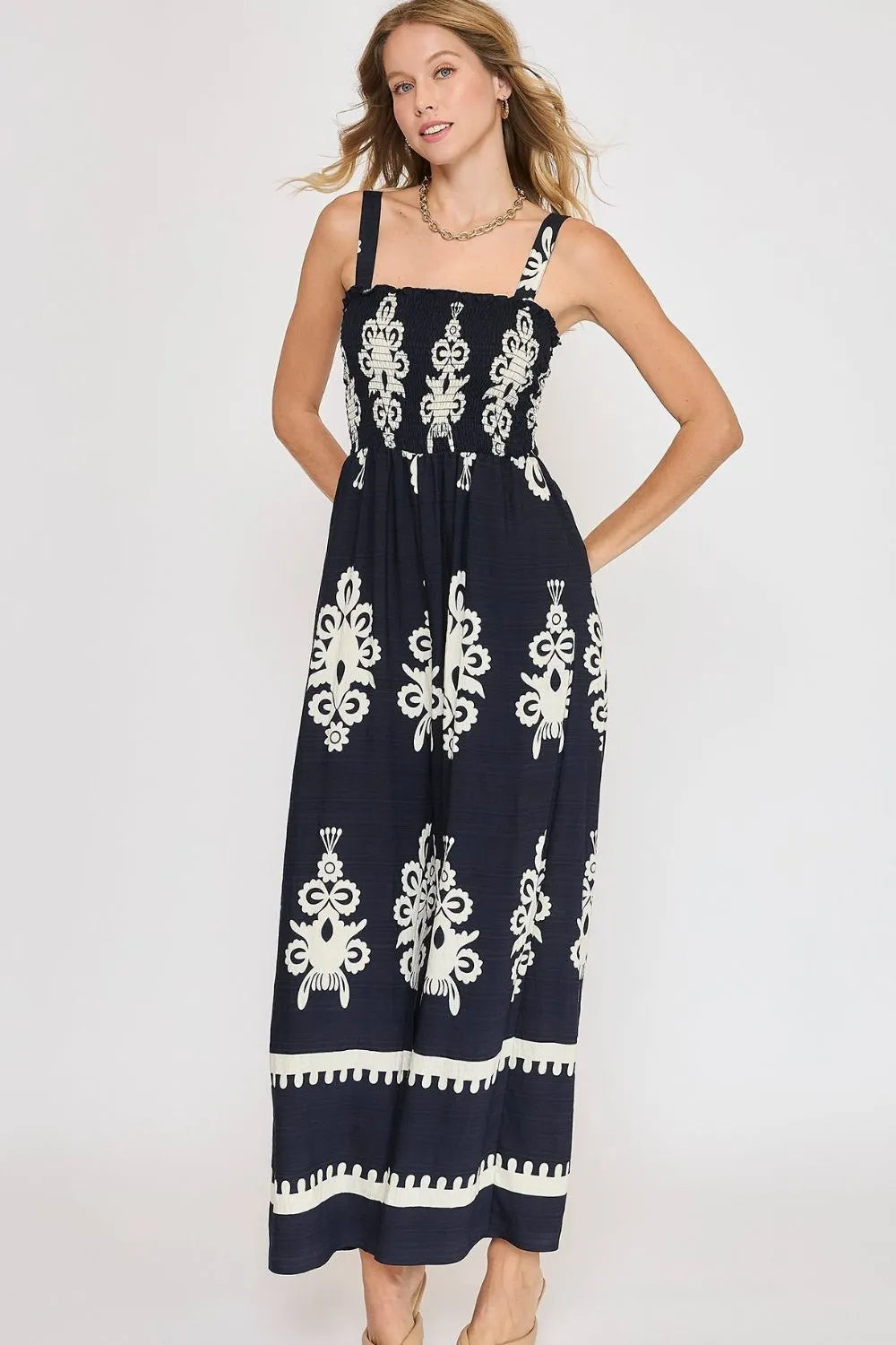 Woman in navy blue maxi dress with white embroidered floral patterns and pom-pom trim details, wearing gold chain necklace, posed against light bac...