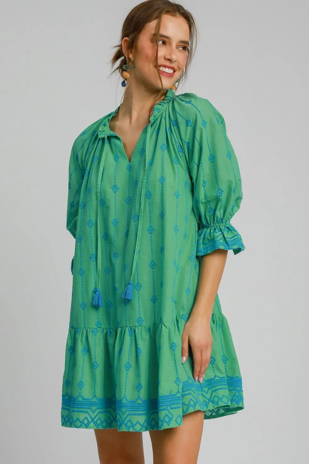 Woman in green dress with blue geometric pattern and puffy sleeves, smiling and posing against white background. Dress features tassel ties at neck...