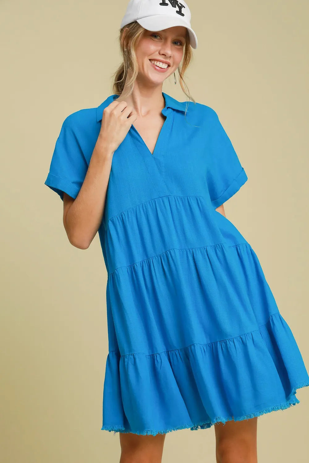 Woman in bright blue tiered dress with short sleeves and frayed hem, wearing white baseball cap, smiling against beige background in casual pose wi...