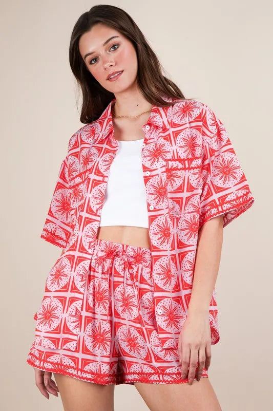 Young woman wearing a red and white tropical print matching shirt and shorts set with a white crop top, posed against a beige background.