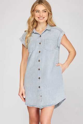 Woman wearing light blue denim shirt dress with short sleeves, button-front closure, and chest pockets, standing with hand on hip against neutral b...