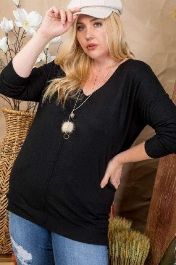 Plus size model wearing black V-neck hi-lo sweater with 3/4 sleeves and side slits, paired with jeans, standing in a home setting.