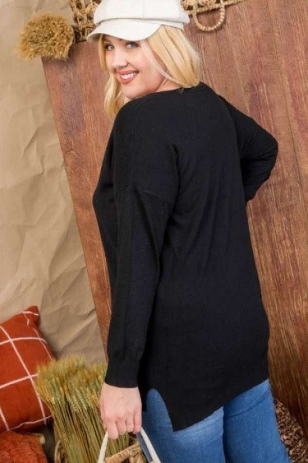 Woman wearing black plus size V-neck hi-lo sweater with 3/4 sleeves, styled with a cap and jeans, standing in cozy room setting.