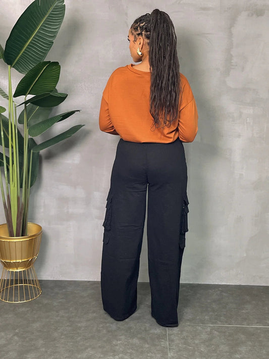 Woman wearing black plus size wide leg cargo pants with utility pockets, styled with an orange top, standing by potted plant.