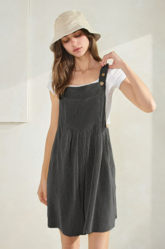 Woman in olive green corduroy overall dress over white t-shirt and beige bucket hat, standing with hand on hip against neutral background.