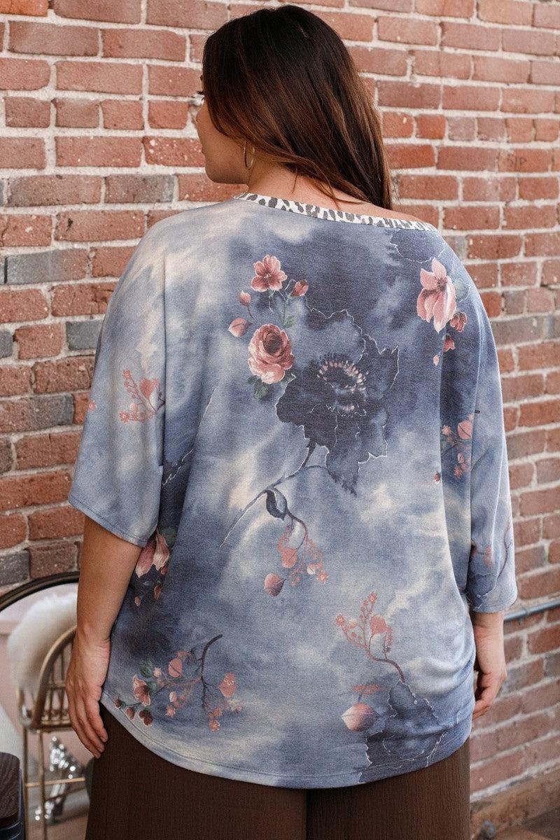 Woman wearing a floral denim print block French terry top with 3/4 dolman sleeves, standing against a brick wall.