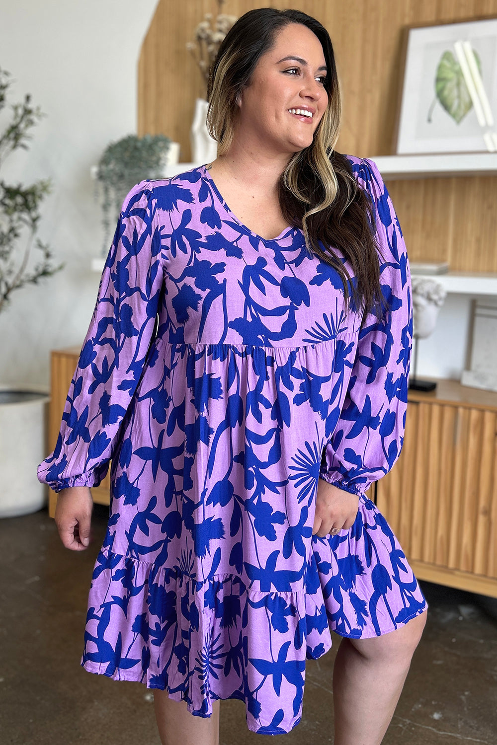 Woman wearing a printed ruffle hem dress with pocket in purple and blue, featuring a floral pattern and long sleeves.