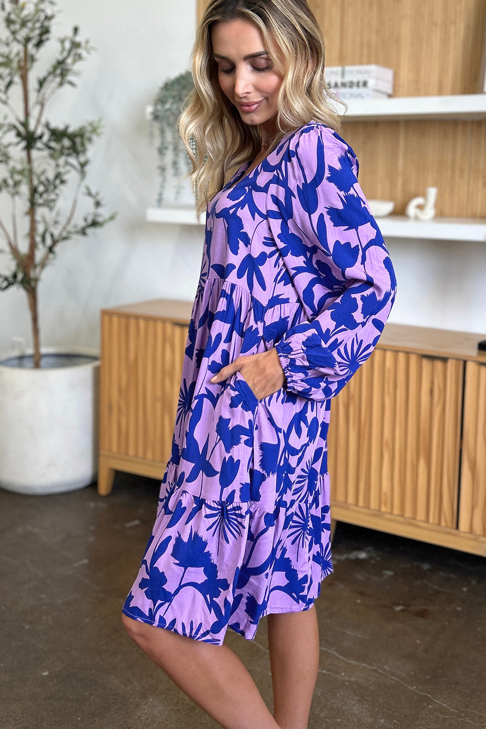 Woman wearing a printed ruffle hem dress with pocket in purple and blue floral design, standing in a stylish living room.