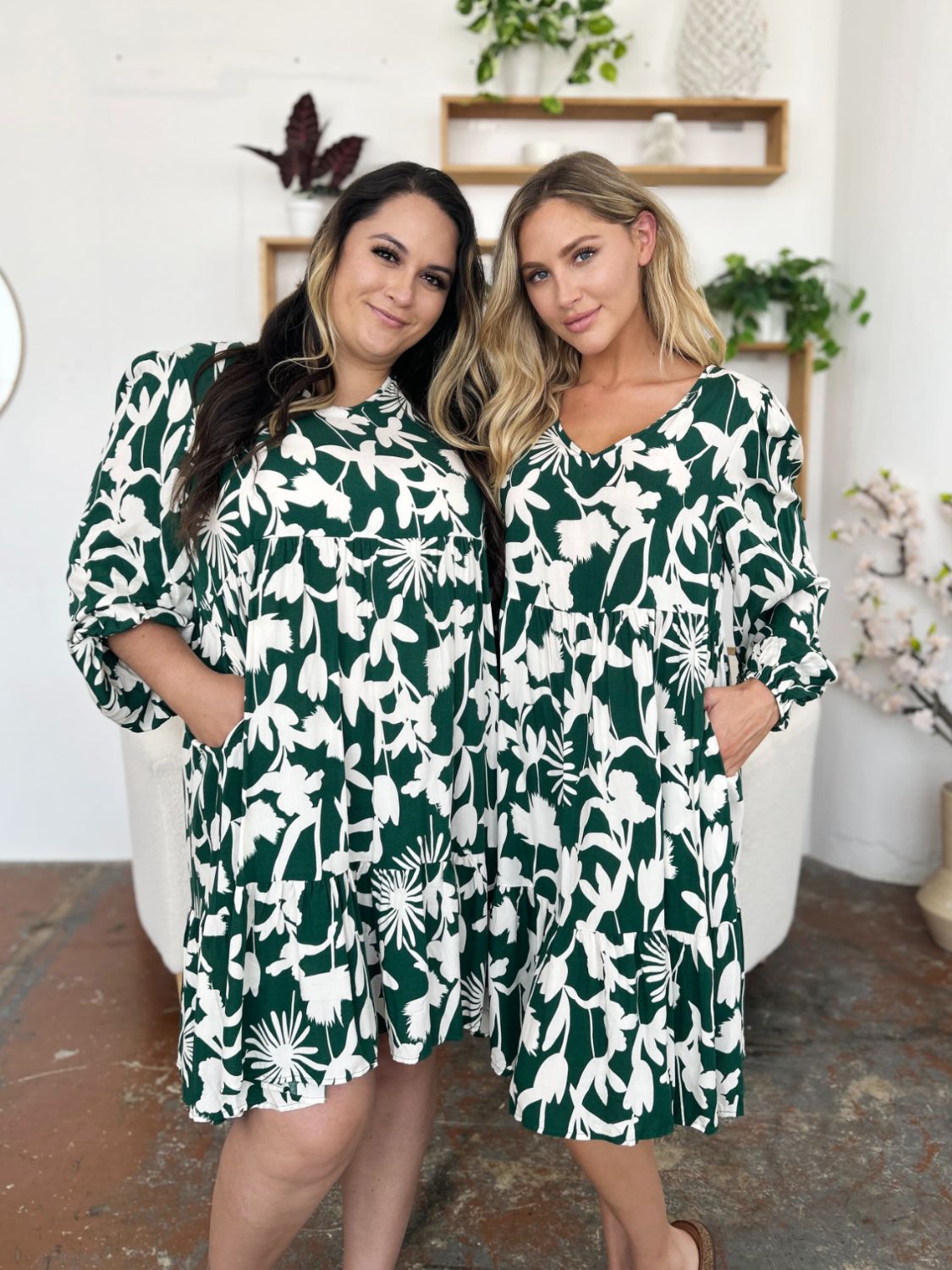 Two women wearing printed ruffle hem dresses with pockets, featuring green floral patterns.