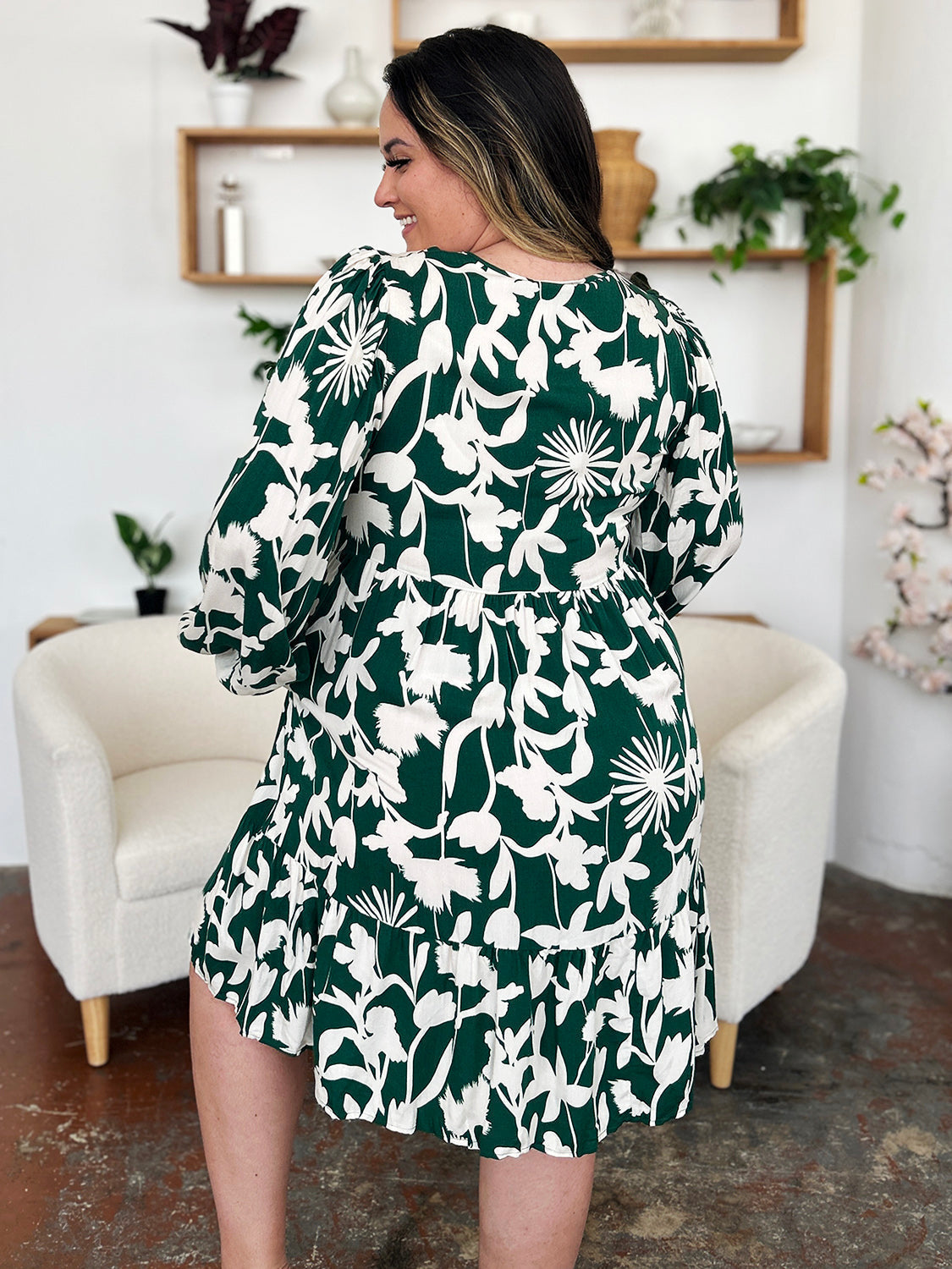 Woman wearing printed ruffle hem dress with pocket, featuring floral pattern, standing in a chic room with white armchairs.