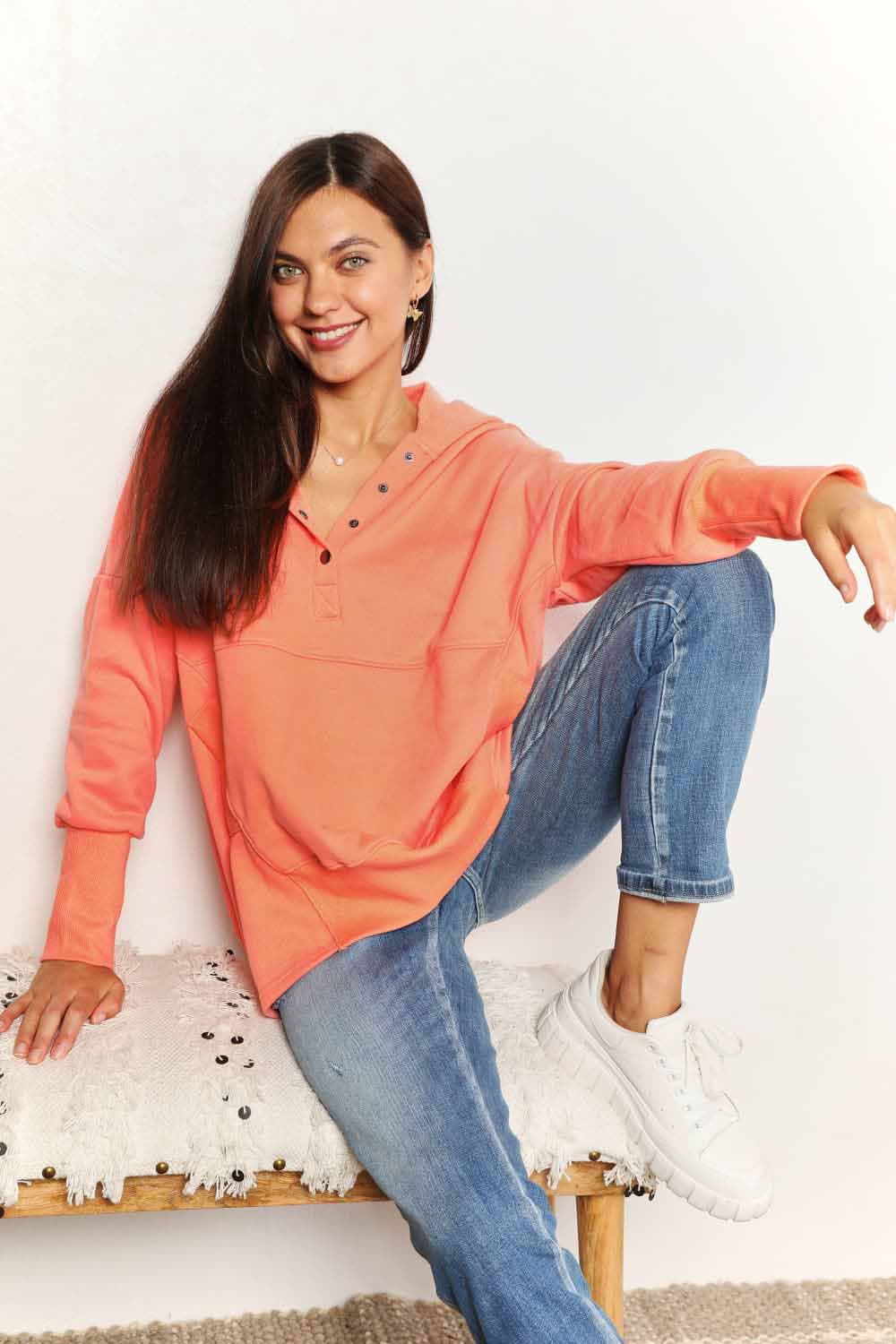 Woman in casual orange quarter-snap dropped shoulder hoodie with pocket, sitting on a bench, paired with jeans and sneakers.