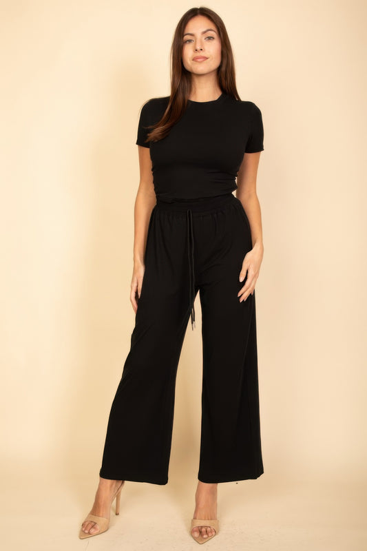 Woman in black short-sleeve top and wide-leg pants with drawstring waist, standing against beige background wearing nude strappy heels.