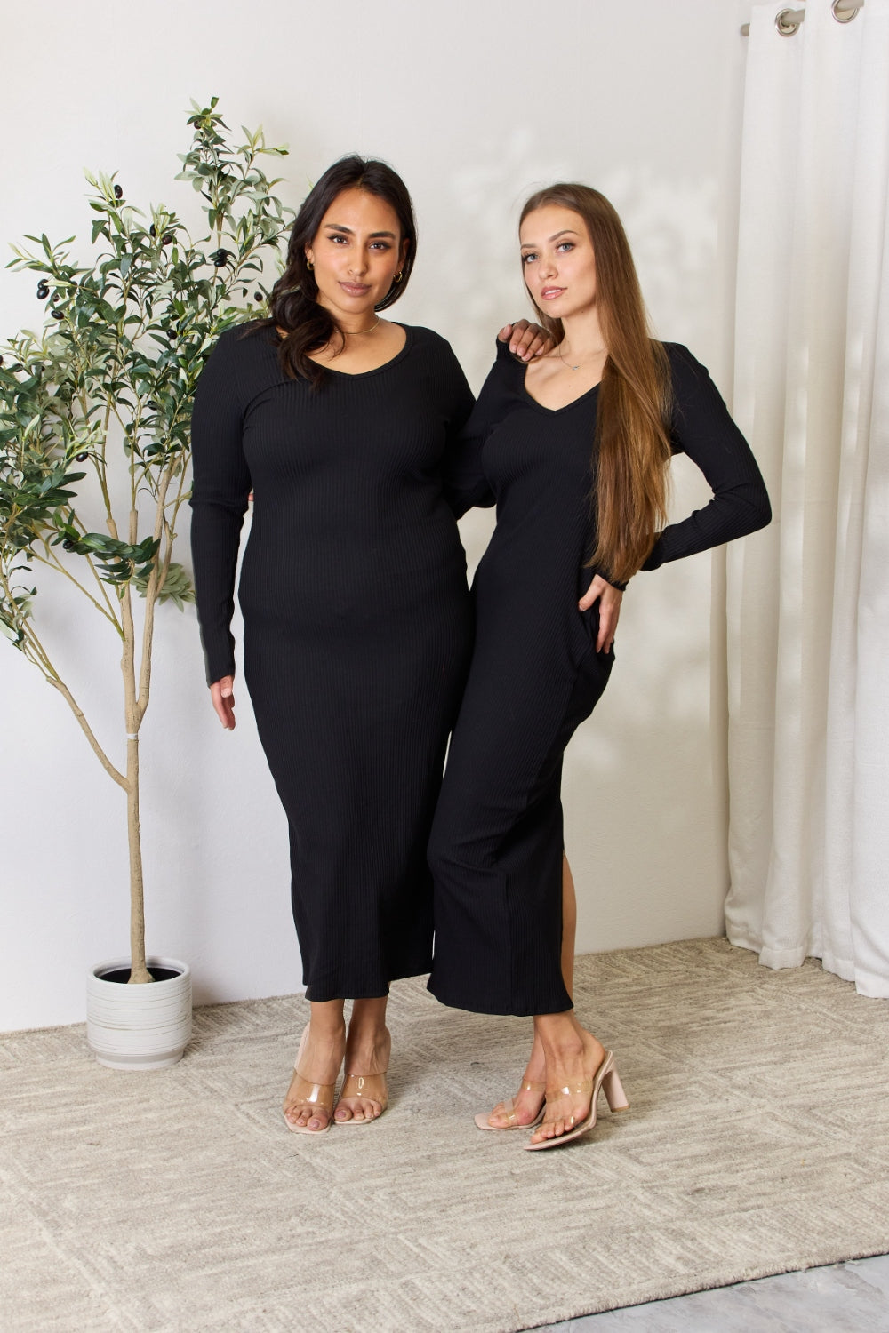 Two women modeling ribbed long sleeve midi slit dresses in black with a moderate stretch, standing on a carpet with a plant background.