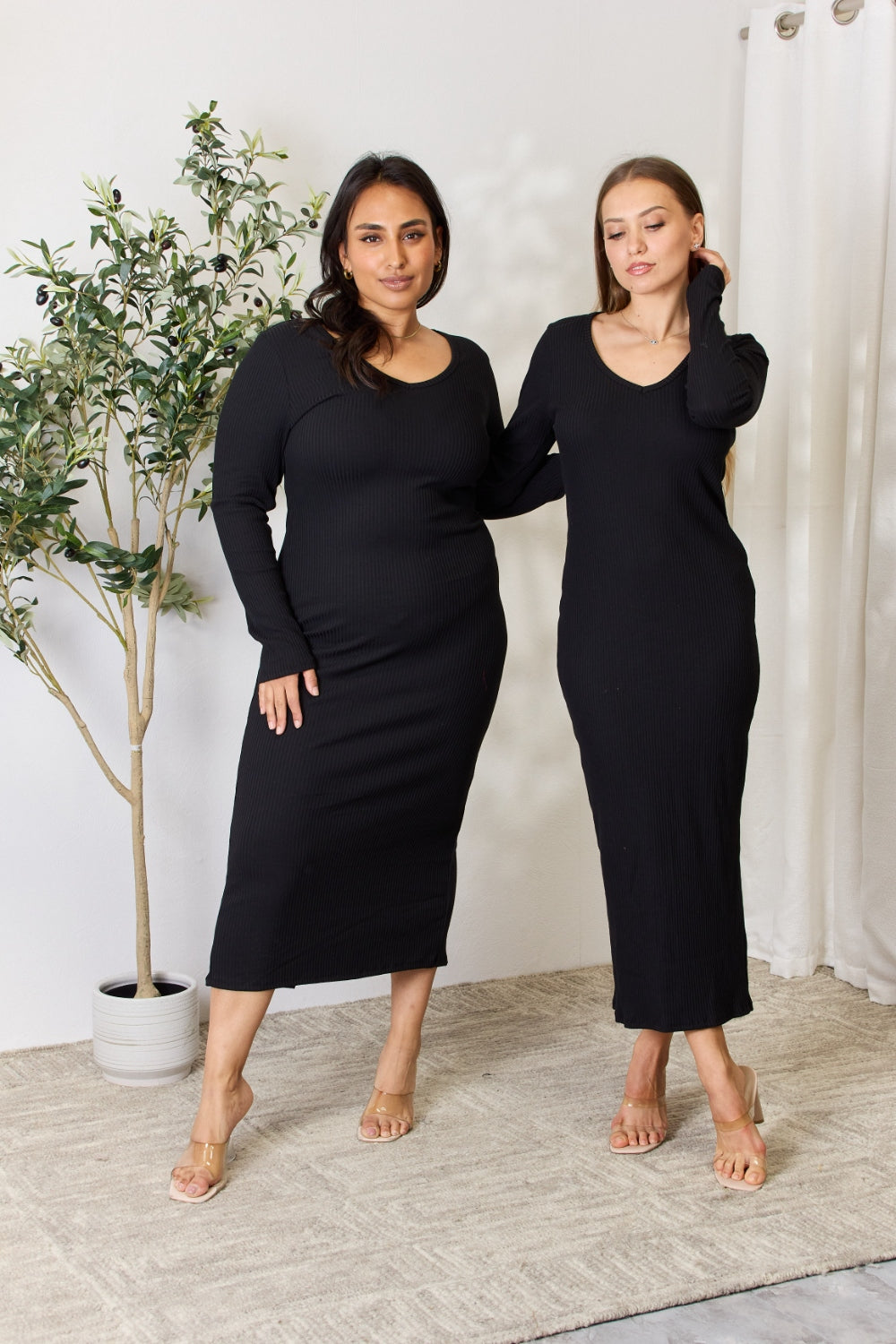 Two women wearing black ribbed long sleeve midi slit dresses, standing together on a beige carpet with a potted plant in the background.