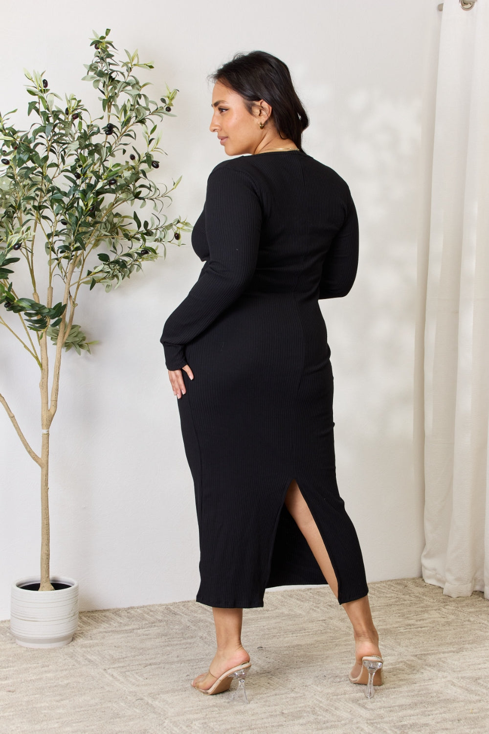 Woman wearing a ribbed long sleeve midi slit dress in black, showing back view with slit detail, standing near a potted plant.