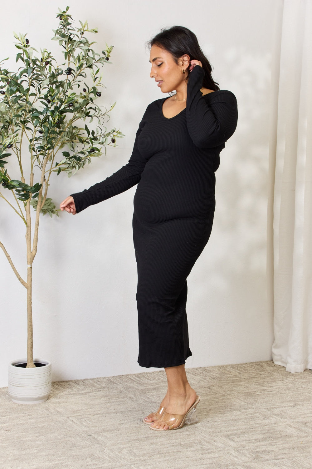 Woman wearing a ribbed long sleeve midi slit dress in black with moderate stretch, standing next to a potted plant in a minimal room.