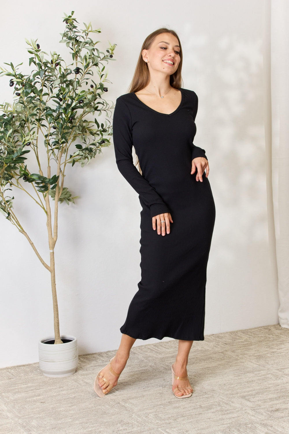 Woman wearing a black ribbed long sleeve midi slit dress standing near a potted plant.