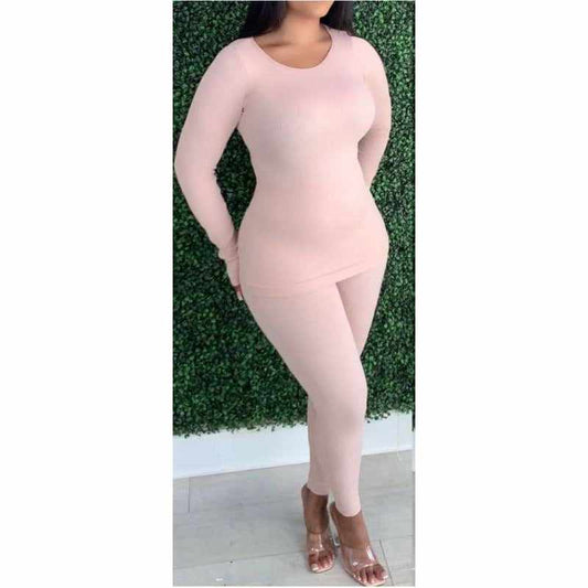 Woman wearing nude pink ribbed lounge two-piece set made of nylon and spandex, standing against a green foliage wall.