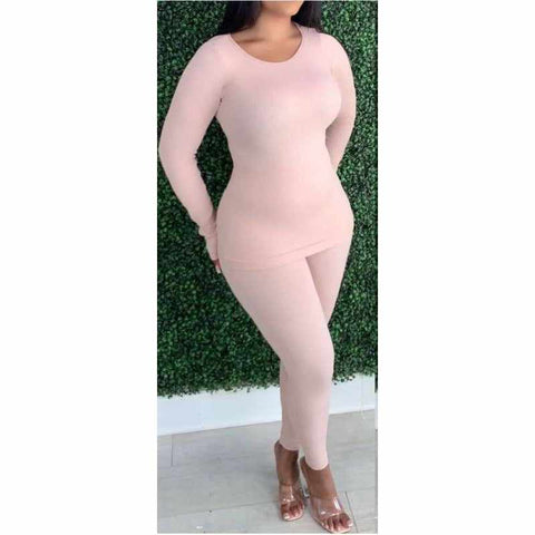 Woman wearing nude pink ribbed lounge two-piece set made of nylon and spandex, standing against a green foliage wall.