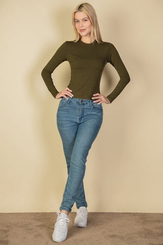 Model wearing a ribbed round neck long sleeve top in olive green, paired with blue jeans and white sneakers, standing on a beige carpet.