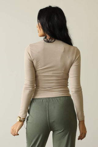 Woman wearing ribbed round neck long sleeve top, beige color, made of polyester and spandex, paired with green pants, rear view.