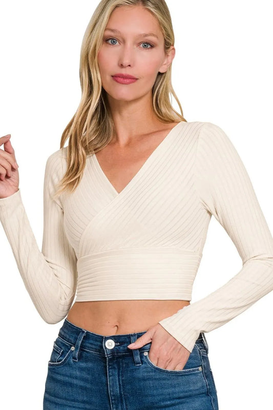 Woman with blonde hair wearing cream-colored ribbed long-sleeve crop top and blue jeans, standing with one hand on hip against white background.