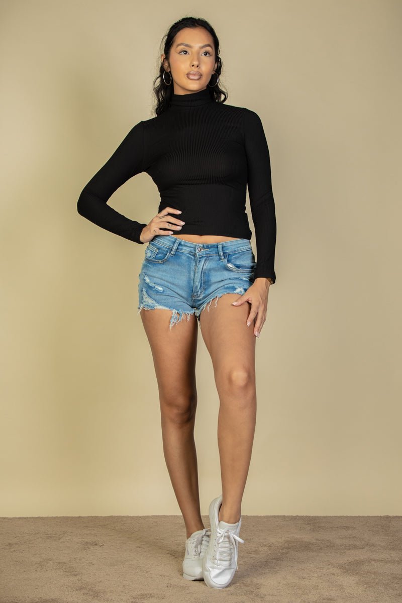 Model wearing black ribbed turtleneck long sleeve top, paired with denim shorts and white sneakers, standing against beige background.