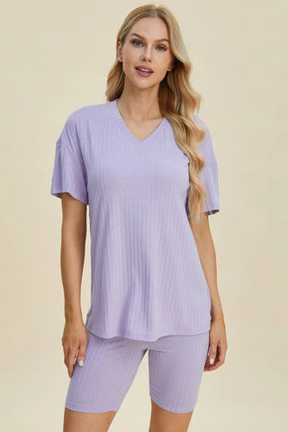 Woman with long blonde hair wearing a lavender purple ribbed short-sleeve v-neck top and matching shorts, standing against a cream background.