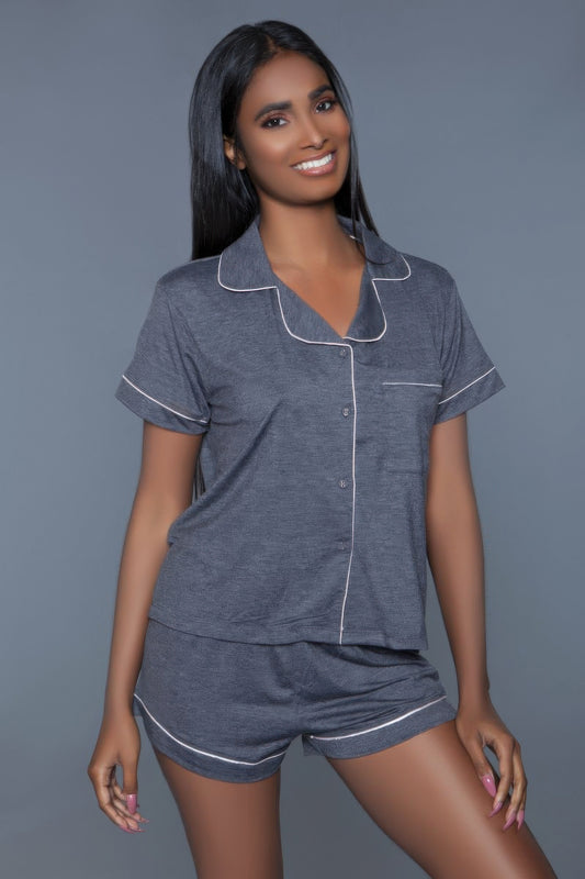 Woman in gray pajama set with white piping trim, smiling at camera against gray background. Short-sleeve button-up top and matching shorts in...