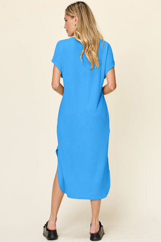 Woman wearing a blue round neck short sleeve slit dress made from slightly stretchy polyester spandex fabric, back view.
