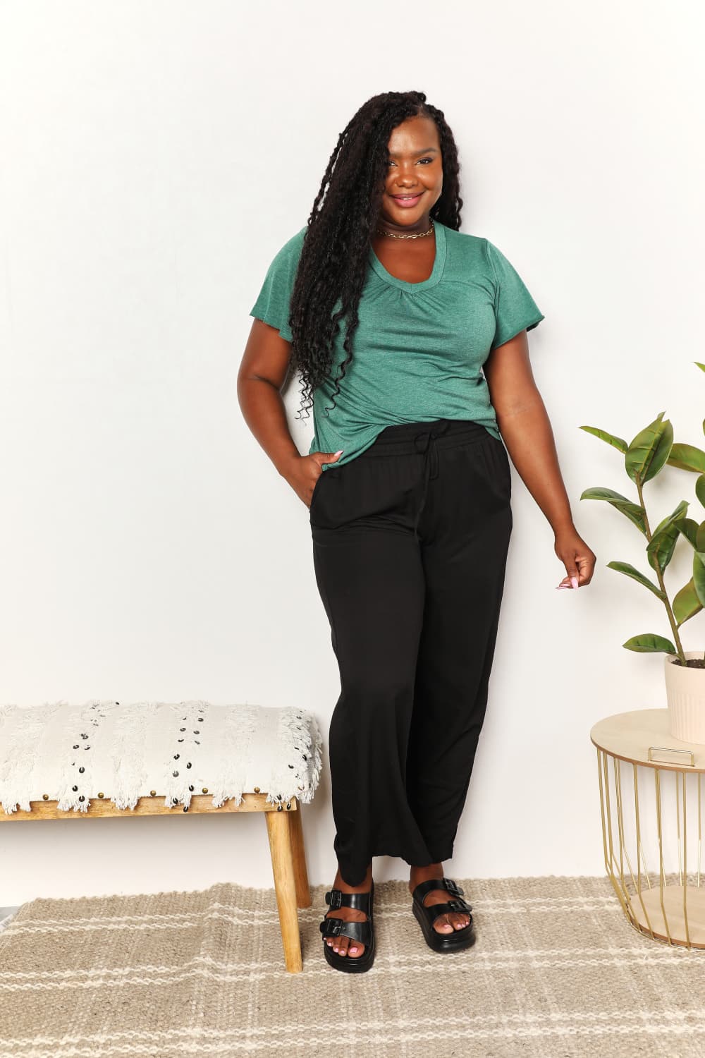 Woman wearing a green ruched V-neck T-shirt and black pants, standing in a stylish room with a plant and a modern bench.