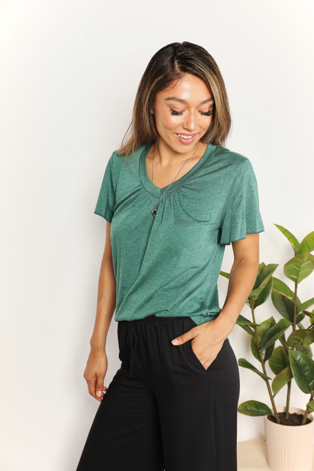 Woman wearing a green ruched V-neck short sleeve T-shirt with black pants, standing beside a plant indoors.