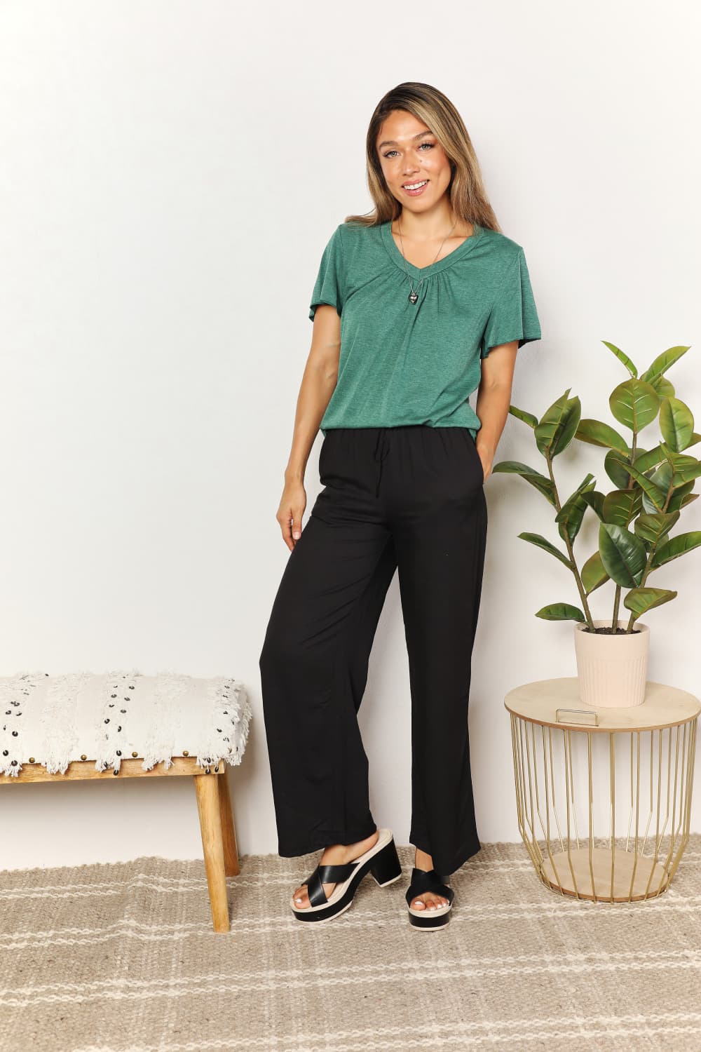 Woman wearing chic green ruched V-neck T-shirt and black pants, standing stylishly beside a plant and bench.