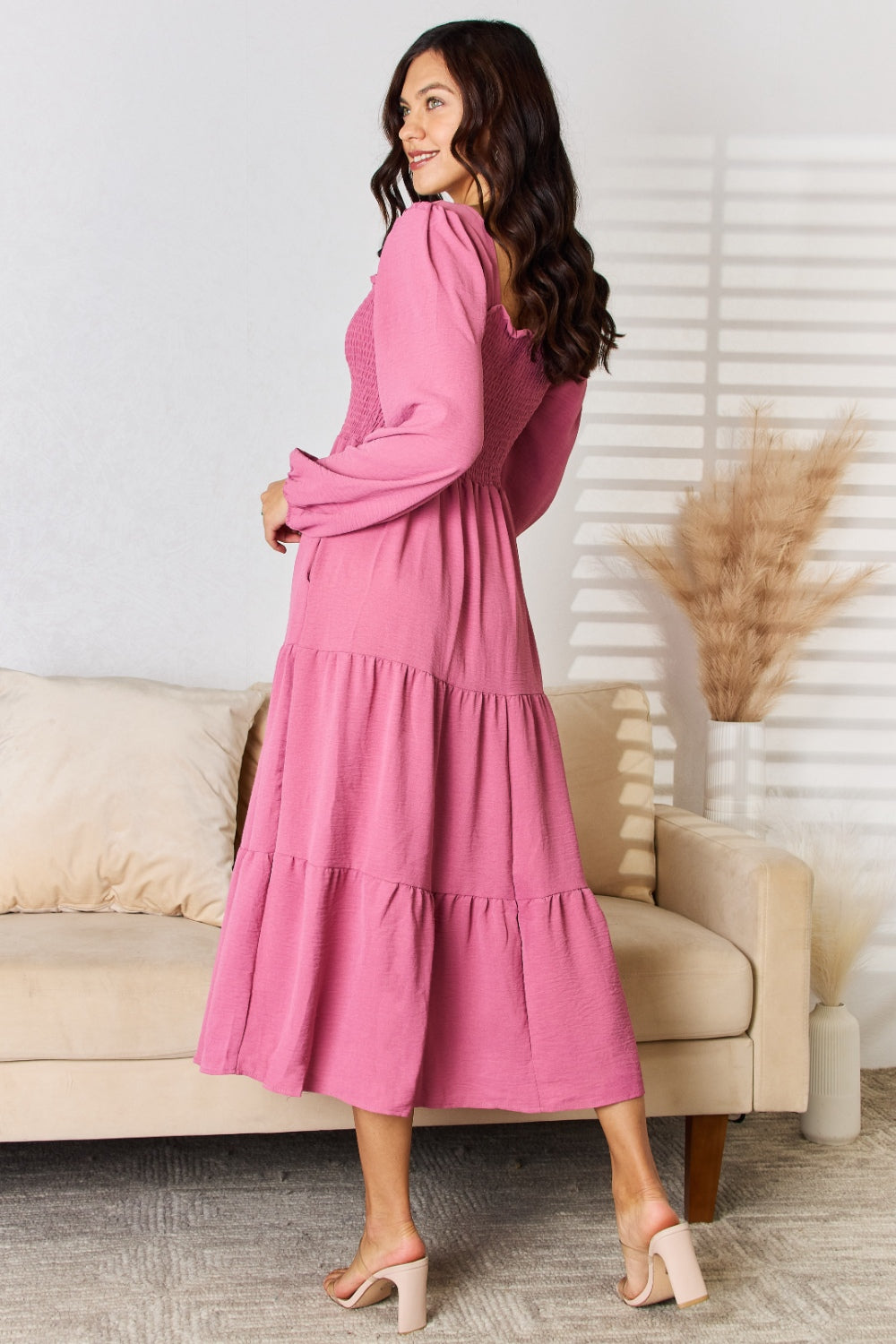 Woman in pink ruffle trim smocked tiered dress standing in stylish room with a sofa and pampas grass.