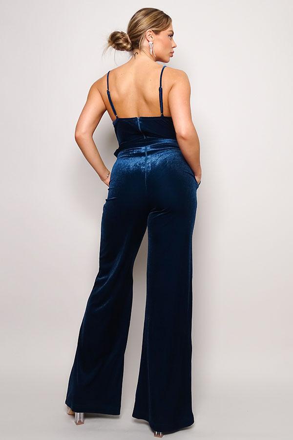 Woman wearing teal velvet jumpsuit with rhinestone belt, showing back view and adjustable straps, featuring long, loose-fitting pants.