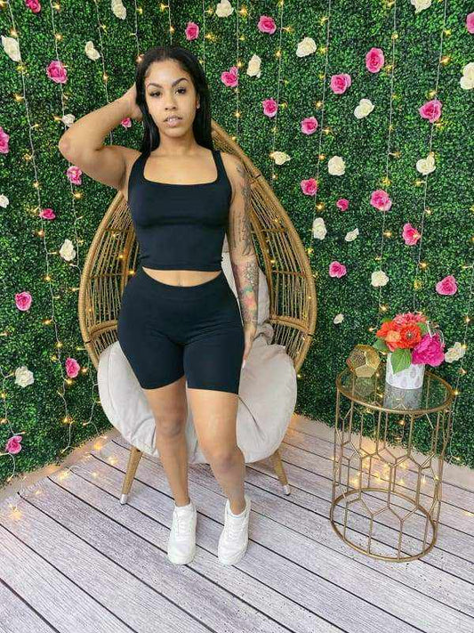 Woman in black seamless scoop neck crop top and biker shorts set, posing against floral wall, wearing white sneakers.