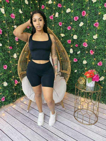 Woman in black seamless scoop neck crop top and biker shorts set, posing against floral wall, wearing white sneakers.