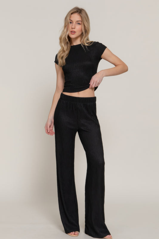 Woman in black ribbed two-piece set with short-sleeve crop top and wide-leg pants, standing with hand on hip against neutral background.