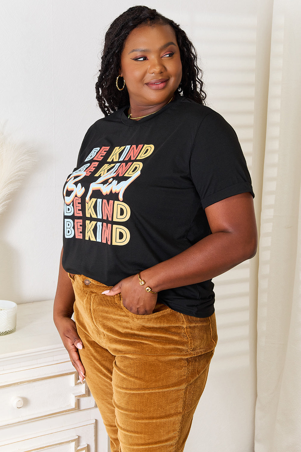 Woman wearing black "BE KIND" graphic round neck t-shirt with mustard pants, standing indoors.