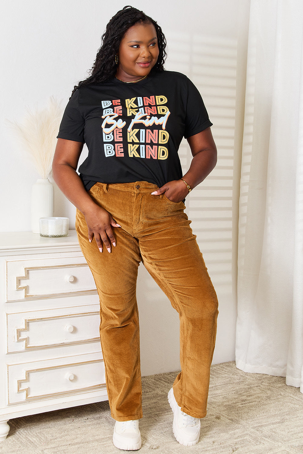 Woman wearing Simply Love BE KIND graphic round neck T-shirt with brown pants, standing in a stylish room.